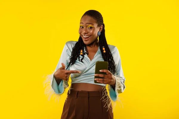 Happy Black Lady Using Cellphone And Pointing Finger At Smartphone Advertising New App Standing On Yellow Studio Background, Smiling To Camera. Try This Mobile Application Concept