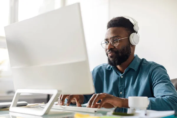 African american male employee in glasses and wireless headphones sitting at office desk and working on computer. Black man in modern headset browsing internet