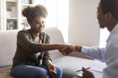 Gratittude for work. Selective focus on cheerful happy young african american woman handshaking with man psychologist after successful therapy session, sitting on couch at counselor office, copy space