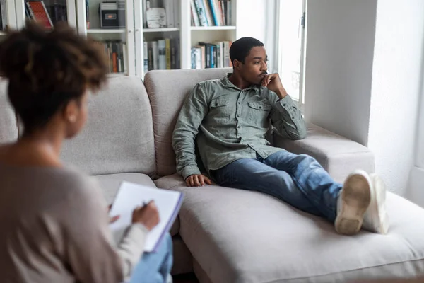 Frustrated pensive young african american man in casual outfit reclining on sofa at psychologist office, having therapy session. Black woman psychotherapist helping depressed guy with PTSD, copy space