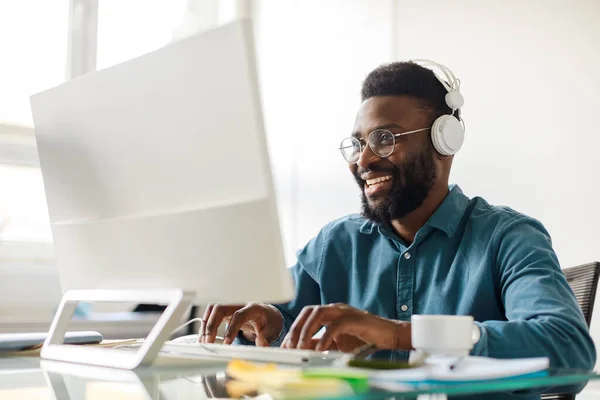 Happy successful black businessman in headset typing on computer at workplace in office interior, consulting client and having video call. Business with modern gadgets
