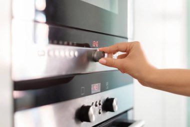 Female hand adjusting temperature on electric oven while cooking in kitchen, unrecognizable woman turning knob, selecting functions regime on modern stove while preparing food at home, cropped