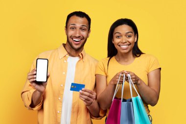 Smiling young african american couple hold packages with purchases, use credit card, show smartphone with blank screen, isolated on yellow background, studio. Cashback app, shopping together and sale