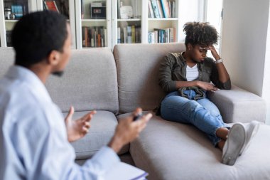 Depressed unhappy young african american woman in casual outfit reclining on couch at psychologist office, having therapy session. Black man psychotherapist helping lady with anxiety, loneliness