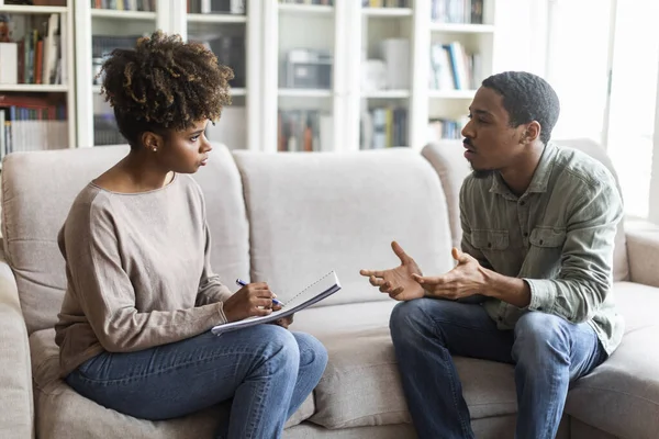 Young african american woman therapist taking notes on her upset patient at therapy session, frustrated black man having conversation with psychologist, sharing feelings, counselor office interior