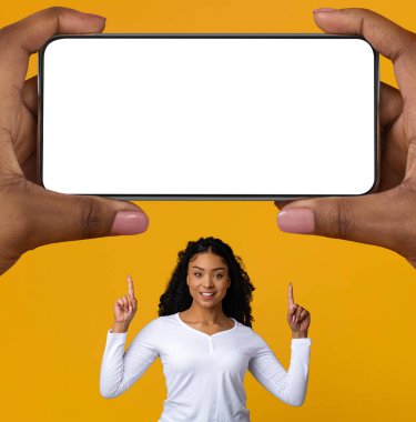 Happy Black Woman Pointing Up At Big Blank Smartphone In Giant Female Hands Over Yellow Background, Smiling Young African American Lady Recommending New Mobile Application Or Online Offer, Mockup