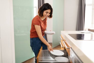Young arab woman placing clean dishes in kitchen drawer after washing, smiling middle eastern female tidying up at home, happy housewife making general cleaning, enjoying domestic chores