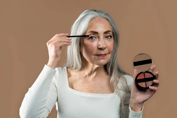 Calm aged european lady with gray hair applies shadows with brush on her eyes, shows cosmetics, isolated on brown background, studio. Ad and offer, anti-aging nude makeup, beauty care, daily procedure