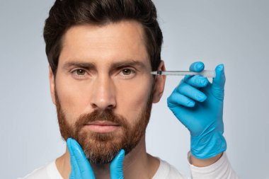 Cosmetologist doctor making botox injection for eyes area to middle aged man, bearded male getting anti-wrinke treatment, standing over grey sudio background