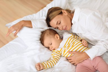 Mother and little baby infant girl lying sleeping together in cozy white bed at home, above view, free space. Loving mom and small newborn daughter taking nap fall asleep. Parenthood concept