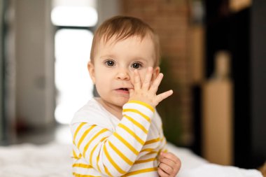 Teething concept. Portrait of little baby girl in bodysuit sitting on white bedsheets and biting finger in mouth, looking at camera. Children and nursery