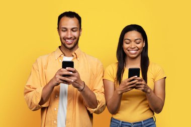 Cheerful young african american husband and wife chatting on smartphones, isolated on yellow background, studio. Relationships and love, gadget addiction, app social networks and blog, ad and offer