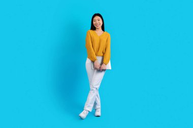Happy attractive bright young asian woman in stylish outfit digital nomad with modern laptop posing over blue studio background, full length, copy space. Freelance, remote job concept