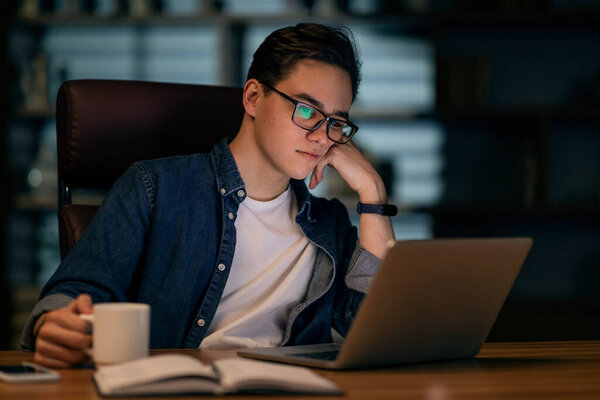 Tired handsome young guy wearing eyeglasses manager looking at laptop screen and touching his face, developer looking for solutions while working at office late at night, drinking coffee, copy space
