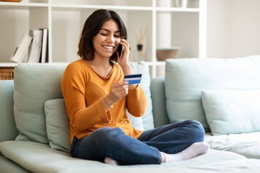 Young Smiling Arab Woman Holding Credit Card And Talking On Mobile Phone At Home, Happy Middle Eastern Female Making Online Purchases While Sitting On Comfortable Couch At Home, Copy Space