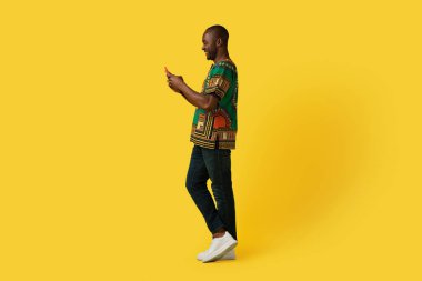 Positive smiling happy mature african american man in traditional costume using modern cell phone over yellow studio background, checking newest mobile app, copy space, full length shot