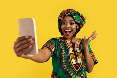 Emotional happy pretty young african woman in traditional clothes and bright makeup looking at smartphone screen and gesturing, celebrating success, won lottery online, yellow studio background