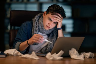 Sick young man in casual wearing warm scarf around neck sitting in front of computer at home office late at night, checking body temperature, holding thermometer, copy space. Cold, flu, coronavirus clipart