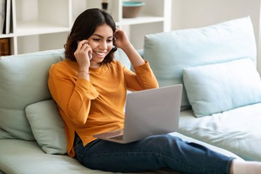 Happy Young Arab Woman Talking On Cellphone And Shopping Online On Laptop At Home, Smiling Middle Eastern Female Making Internet Purchases Via Computer And Enjoying Phone Call, Free Space