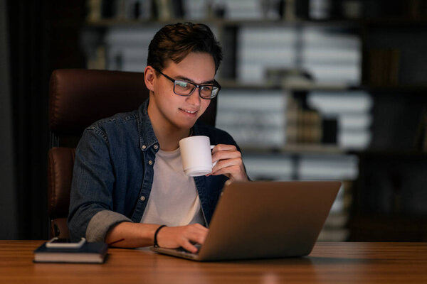 Overworked cheerful handsome young man in casual wearing eyeglasses typing on laptop keyboard and holding white cup, drinking coffee while working late at night at office, copy space