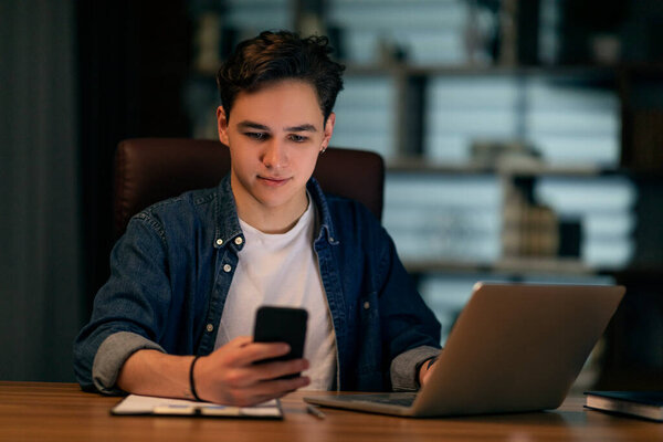 Young handsome guy in casual manager working late at office, sitting at workdesk, using modern laptop and smartphone, working on monthly report, using business mobile app, copy space