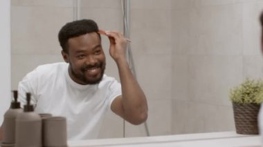 Young african american guy preening himself in morning, combing his short hair and smiling, looking at mirror at bathroom, tracking shot, slow motion, free space