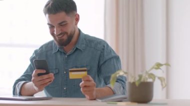Convenient banking service. Young positive middle eastern guy making online payment on smartphone, using credit card and mobile application at home, tracking shot, slow motion, empty space