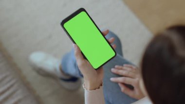 Online order. Top view of unrecognizable woman holding smartphone with green chroma key screen, studying assortment on mobile app, choosing product, empty space