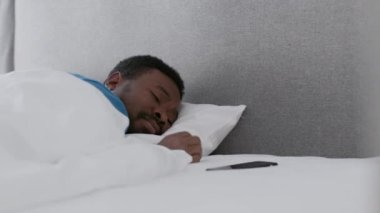 Annoying spam. Young black guy sleeping in bed and waking up because of message notification on smartphone, turning away and keep resting, slow motion, empty space
