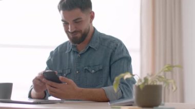 Online communication concept. Young positive arab guy web surfing on smartphone, chatting with friends and reading news, resting at workplace, tracking shot, slow motion, empty space