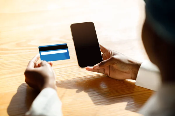 Unrecognizable black woman customer holding modern smartphone with black empty screen and blue credit card, checking CVV number, female shopping or banking online, mockup, cropped shot