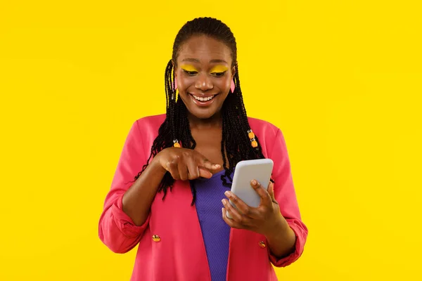 Excited Black Woman Using Smartphone Pointing Finger Choosing Mobile Application Standing Over Yellow Studio Background. Female Posing With Phone Advertising New App