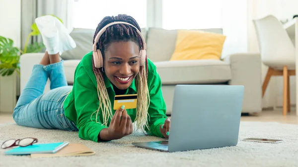Online shopping. Positive african american woman showing credit card to laptop webcam, making payment, lying on floor at home, panorama. Internet commerce and banking service concept