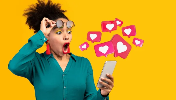 Happy surprised young black lady with open mouth has romantic chat with hearts on phone enjoys message isolated on orange studio background, close up, panorama. App for dating, relationships and love
