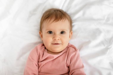 Adorable caucasian baby in bodysuit lying on bed at home, resting on white bedsheets in bedroom, closeup portrait, free space, top view