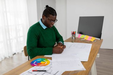 African American Male Designer Working Drawing A Plan Using Color Palette Sitting Near PC Computer In Modern Office, Wearing Eyeglasses. Successful Web Design Career Concept. Side View Shot