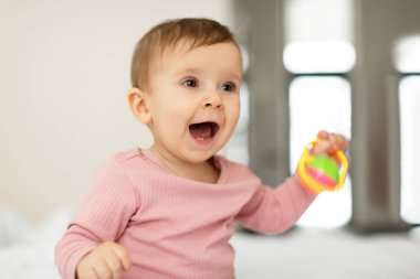 Child care concept. Portrait of pretty little girl sitting in bed with toy and smiling, adorable baby in bodysuit looking away at free space, sweet child playing in bedroom at home
