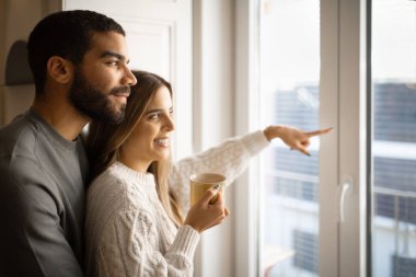 Happy young arab man hug european wife, lady pointing finger out the window and drink cup of tea, enjoy great view and weather at weekend in home interior. Love, relationship, coffee break together