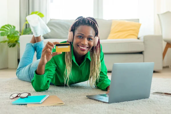 Happy black female buyer holding credit card and using laptop computer, wearing headphones and buying something in internet from home. E-commerce, internet banking