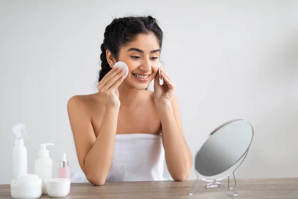 stock image Portrait Of Beautiful Young Indian Woman Cleansing Skin With Cotton Pad Near Mirror, Smiling Millennial Hindu Female Looking At Her Reflection, Enjoying Beauty Skincare Routine At Home, Copy Space