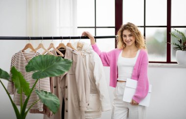 Creative Professions Concept. Smiling Fashion Designer Woman Posing Near Clothing Rail In Modern Showroom, Beautiful Happy Female Stylist Presenting New Clothes Collection And Looking At Camera