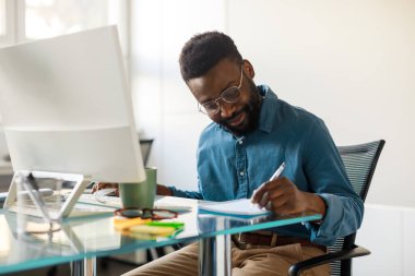 Happy black executive ceo manager using laptop and writing notes in notebook, sitting at workplace in office. Businessman analyzing financial data doing research