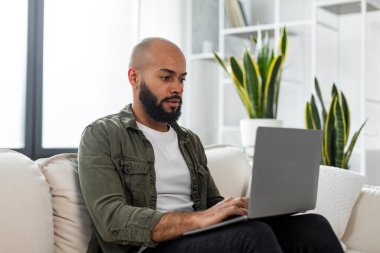 Focused male freelancer working from home, sitting on couch and using modern laptop, typing on keyboard or communicating with clients online, copy space