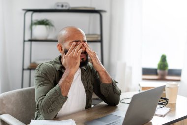 Tired black male rubbing eyes, suffering from overwork, discomfort long hours of sedentary in home office interior. Work, business and health problems clipart