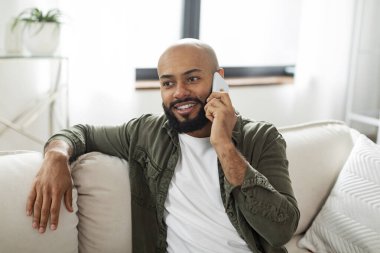Contented latin man talking on cellphone and smiling while sitting on sofa at home, free space. Male communicating by phone, having pleasant conversation