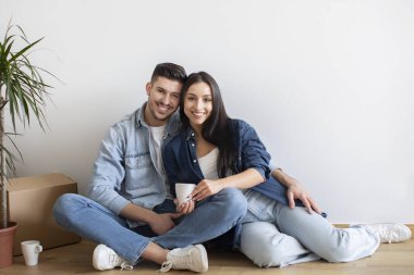 Beautiful Young Couple Relaxing On Floor After Relocation To New Home, Happy Millennial Spouses Embracing And Drinking Coffee Together, Resting Among Cardboard Boxes On Moving Day, Copy Space