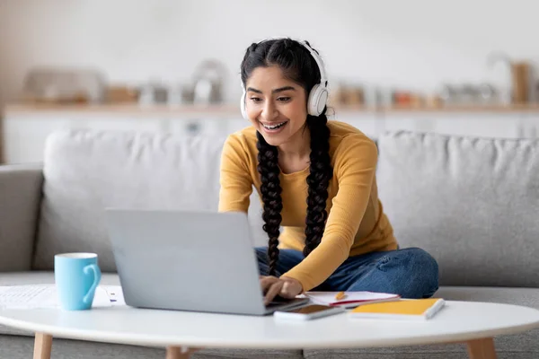 Remote Education. Young Indian Woman In Headphones Studying With Laptop At Home, Smiling Hindu Female Student Using Computer For Distance Learning While Sitting On Couch In Living Room, Free Space