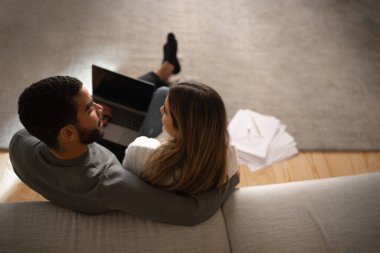 Young arabic male hug and look at european woman typing on computer with blank screen in living room interior with documents and charts. Business, work remotely together, love, relationship at home