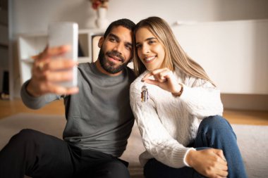 Happy young arab man hug european wife take photo on smartphone and show keys in living room interior. Love, relationship, selfie for blog and social networks, video call at home, buy own apartment