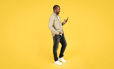 Glad middle aged black man in casual typing on phone, chatting in social networks, surfing in internet, isolated on orange background, studio. App for blog, gadget for communication, work remotely